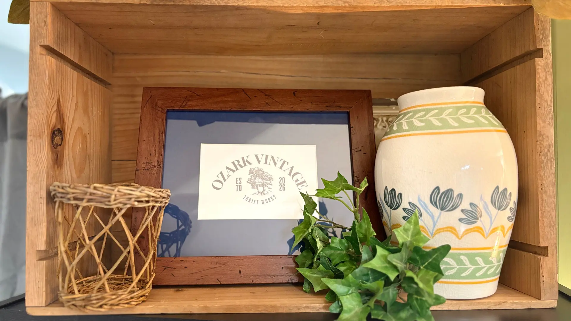 A wooden shelf display with a framed Ozark Vintage Thrift Works sign, a wicker basket, a painted ceramic vase, and trailing ivy