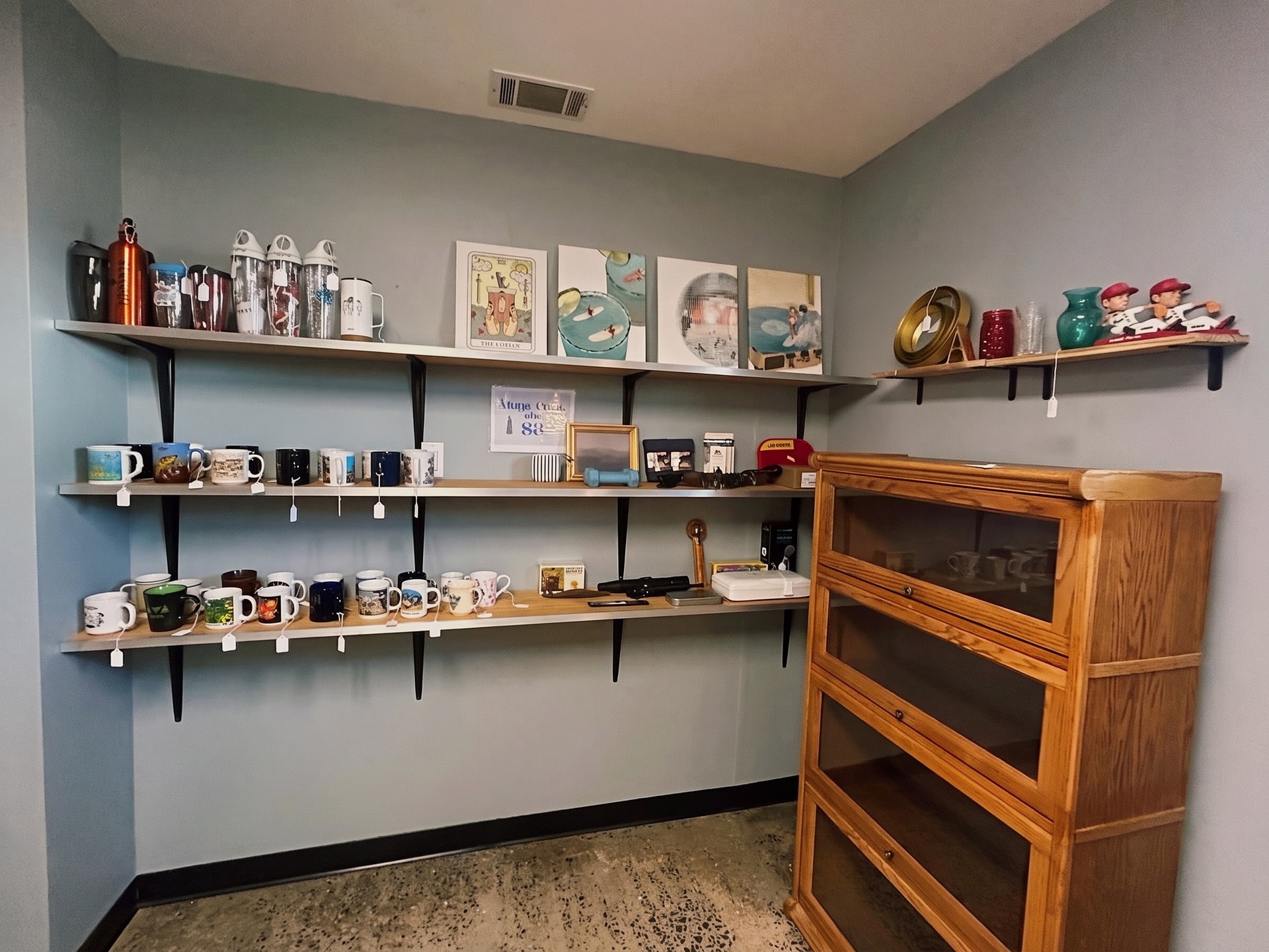 Wall shelves of vintage mugs, tumblers, and art prints beside an oak barrister cabinet